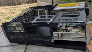 An IBM 5170 chassis with the cover off on a patio table: front panel with keylock, a 5.25 inch floppy drive, the drive cage, a memory card and the power supply