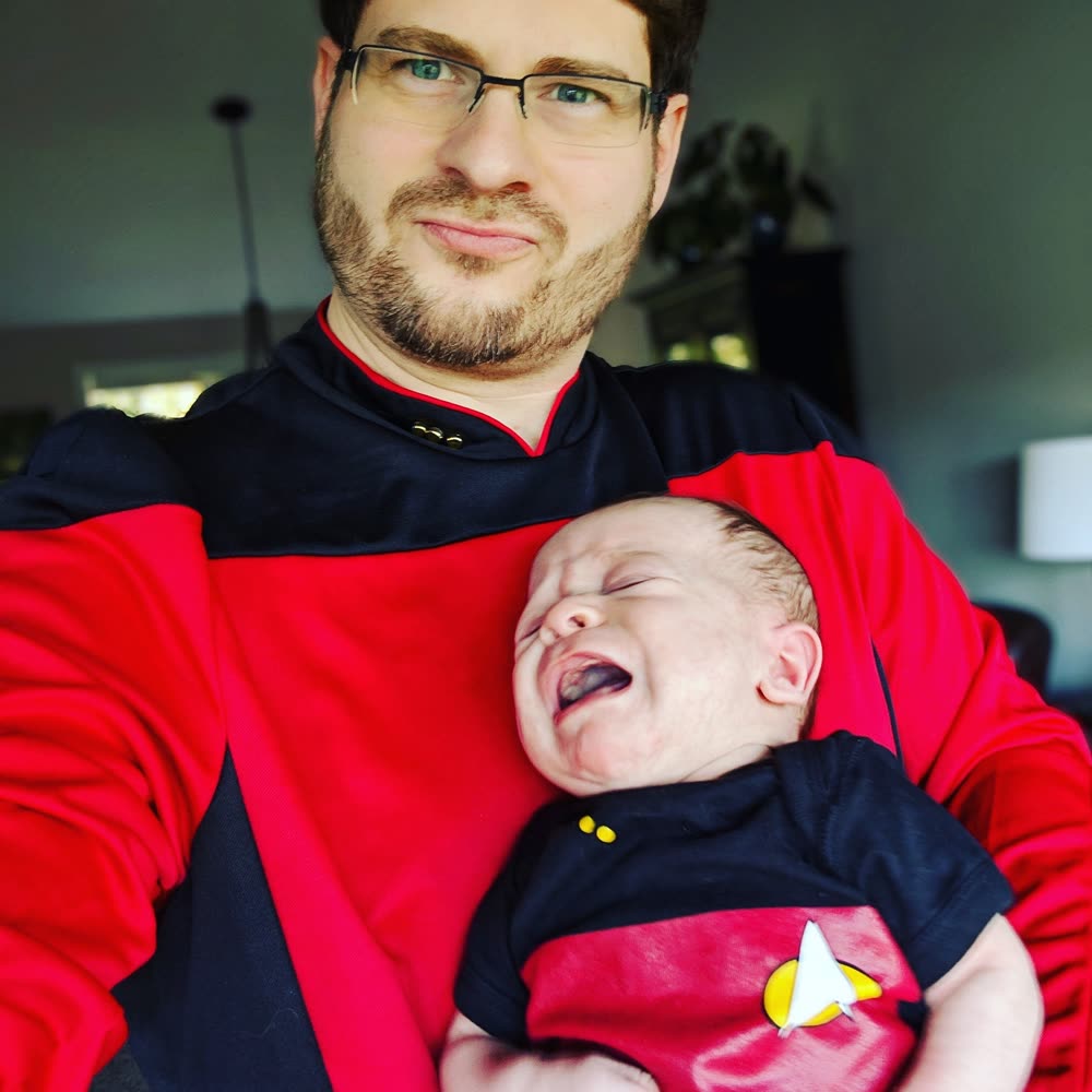 Tony in a Star Trek: The Next Generation command-red uniform, holding a newborn dressed in a matching command-red onesie complete with Starfleet combadge. The baby is mid-yawn. Indoor home setting.